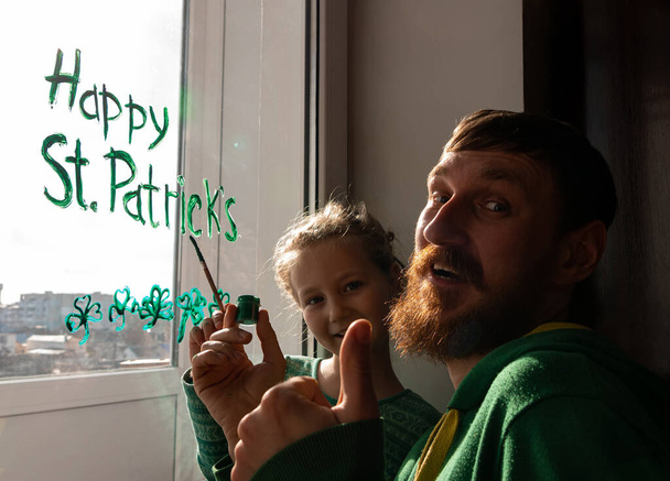 Dessin du père de la Saint-Patrick avec sa fille peignant des trèfles verts à trois feuilles à l'intérieur, décoration de fête, loisirs en famille. Dessin feuilles de trèfle sur verre de fenêtre. Rester à la maison concept Nouveau normal - Photo, image