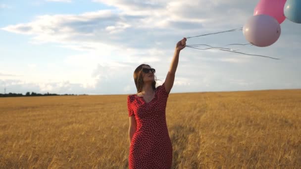 黄金の小麦畑を歩いて手に風船を持って茶色の髪を持つ幸せな若い女の子。大麦のプランテーションで時間を過ごす赤いドレスの美しい女性。自由の概念。スローモーションを閉じます - 映像、動画