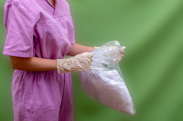 Νεαρή γυναίκα γιατρός σε ροζ ιατρική στολή ξεπακετάρει μια σειρά από ιατρική προστατευτική αντι-πανούκλα κοστούμι. Ο γιατρός φοράει προστατευτική στολή πάνω από τα ρούχα του και ετοιμάζεται να δουλέψει με άρρωστους ασθενείς. Πράσινο πίσω - Φωτογραφία, εικόνα