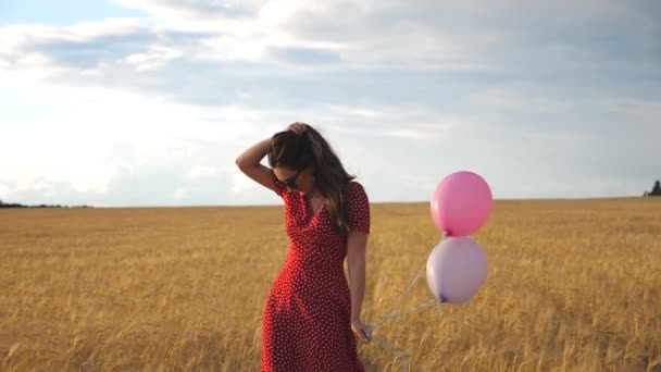 Young woman in sunglasses standing against the background of wheat field and holding balloons in hand. Beautiful girl in red dress straightening her long brown hair in barley plantation. Slow motion - Footage, Video
