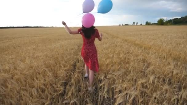 Chica feliz en vestido rojo corriendo por el campo de trigo dorado con globos en la mano en el día nublado. Mujer joven con cabello castaño divirtiéndose mientras trota entre la plantación de cebada. Concepto de libertad - Metraje, vídeo