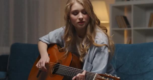 mujer emocional está tocando la guitarra en casa por la noche, retrato medio de la bella dama con instrumento musical - Imágenes, Vídeo
