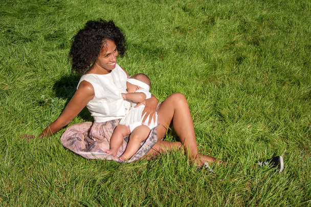 アフリカ系アメリカ人の若い女性が夏の公園で草の上に座って赤ちゃんを母乳で育てている。乳児,母親,授乳,栄養と屋外母乳育児の概念.黒い母親の母乳育児彼女の子供 - 写真・画像