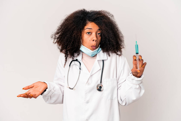 Mladý africký americký kudrnatý lékař žena drží stříkačku pokrčí rameny a otevřené oči zmatený. - Fotografie, Obrázek