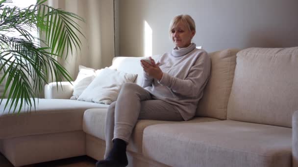 pięćdziesiąt lat kobieta siedzieć na kanapie i używać smartfona - Materiał filmowy, wideo