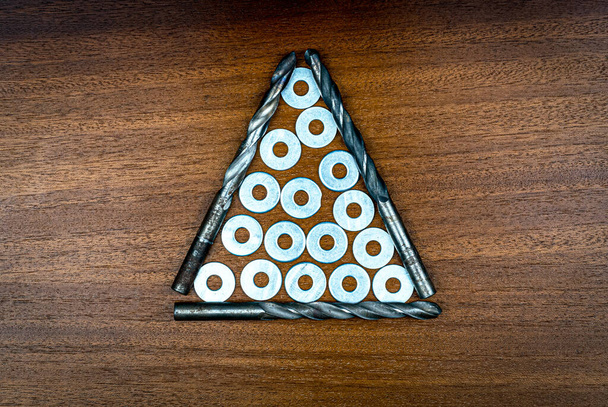 Металлические сверла и шайбы в середине дисплея в виде треугольника на фоне коричневой текстуры дерева - Фото, изображение