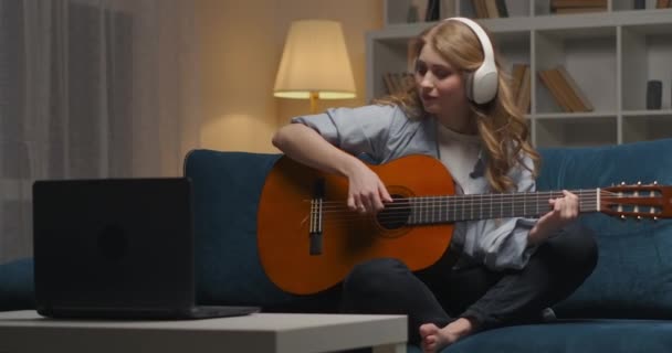 opiskelija tyttö soittaa kitaraa illalla kotona, kuuntelee musiikkia kuulokkeilla ja katselee muistiinpanoja kannettavassa tietokoneessa - Materiaali, video