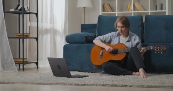 όμορφη κυρία κάθεται στο πάτωμα στο διαμέρισμα και παίζει τραγούδι στην κιθάρα, μάθηση από το διαδίκτυο, μακρινό μάθημα μουσικής - Πλάνα, βίντεο