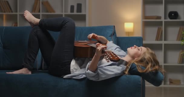 Pensive έφηβος κορίτσι παίζει κιθάρα που βρίσκεται στον καναπέ στο σαλόνι και αισθάνεται λυπημένος, κάνοντας μουσική, θλίψη και μοναξιά - Πλάνα, βίντεο