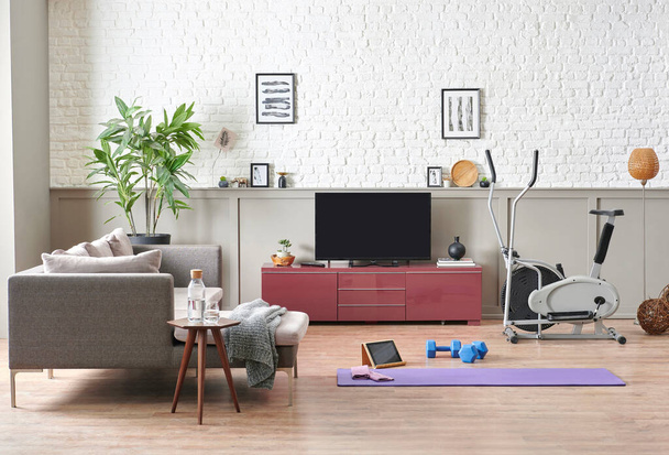 Sportive and training decoration interior room, nobody, television unit, grey sofa and blanket. Purple mat and blue dumbbell on the parquet, white brick wall background. - Photo, Image