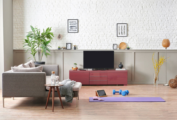Sportive and training decoration interior room, nobody, television unit, grey sofa and blanket. Purple mat and blue dumbbell on the parquet, white brick wall background. - Photo, Image