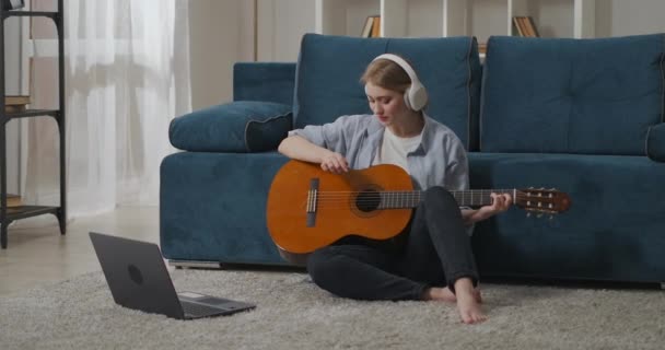νεαρή φοιτήτρια μαθαίνει να παίζει κιθάρα στο σπίτι, βλέποντας φροντιστήριο στο laptop και ακούγοντας μέσω ακουστικών - Πλάνα, βίντεο