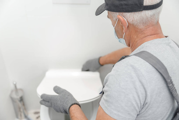 Klempner in medizinischer Maske installiert Toilettenschüssel in Toilette, Arbeit im Badezimmer - Foto, Bild