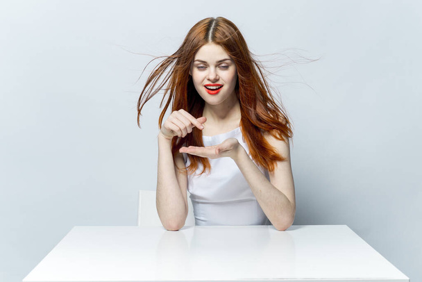 mooie roodharige vrouw zitten aan de tafel gebaar handen emoties en rode lippen model - Foto, afbeelding