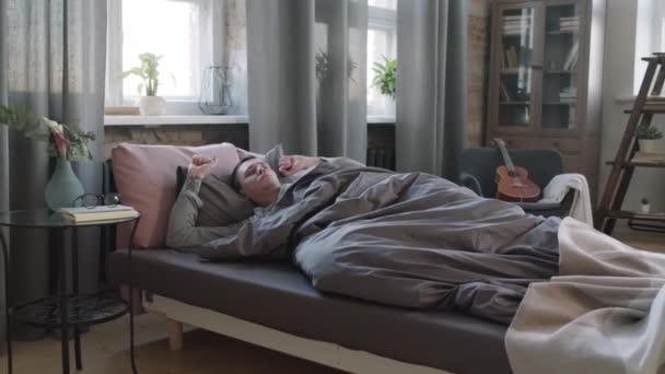 Hidasliikkeinen täysi laukaus unelias mies herää myöhään aamulla, ottaen silmälasit yöpöydästä ja venyttely sängyssä laiskasti - Materiaali, video