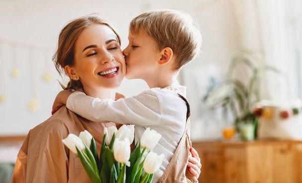 tender son kisses the happy mother and gives her a bouquet of tulips, congratulating her on mother's day during holiday celebration at home - Foto, Imagen