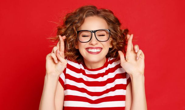 Cheerful young woman in glasses closing eyes and crossing fingers while making wish against red background - Φωτογραφία, εικόνα