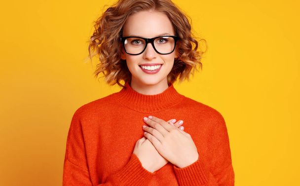 Grateful young woman in orange sweater and glasses holding hands on chest and smiling for camera against yellow background - Photo, Image