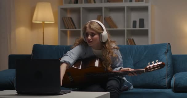 молодая женщина учится играть на гитаре по интернету, включение видео учебник на экране ноутбука, сидя дома вечером - Кадры, видео