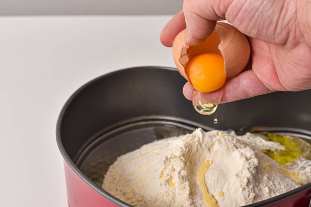 Mano masculina rompe el huevo en harina mientras se hace la masa. Primer plano de la mano del chef con yema de huevo amarillo en proceso de preparación para hornear. - Foto, imagen