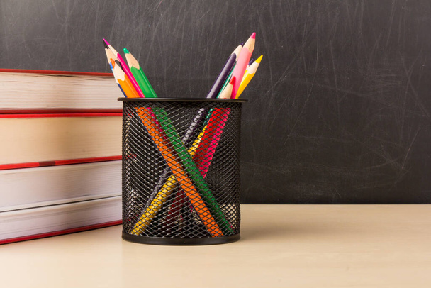 Lápices coloridos en taza de malla de alambre, libros en mesa de madera con pizarra en el fondo, educación o volver al concepto de la escuela - Foto, Imagen