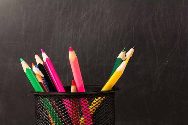 Kolorowe ołówki w czarnym uchwycie. Materiały szkolne i biurowe na stole klasowym przed tablicą. Widok z miejsca kopiowania - Zdjęcie, obraz