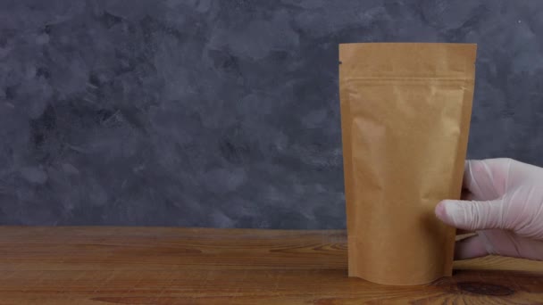 Hand in protective gloves holds brown kraft paper pouch bag with coffee beans wooden background 4K mockup. Packaging for food delivery goods template. Pack with clasp window tea leaves weight product. - Footage, Video
