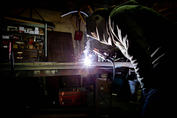 Un soudeur soude des tuyaux métalliques dans un vieil atelier. Le travailleur a un masque de soudure protecteur sur la tête. - Photo, image