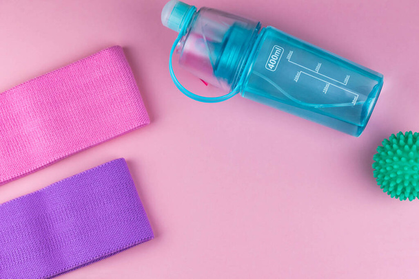 Colorful fitness gums on pink background. Elastic expanders and tapes of different color. Colored rubber and textile bands, bottle of water and massage myofascial ball. Home fitness equipment. - Photo, Image