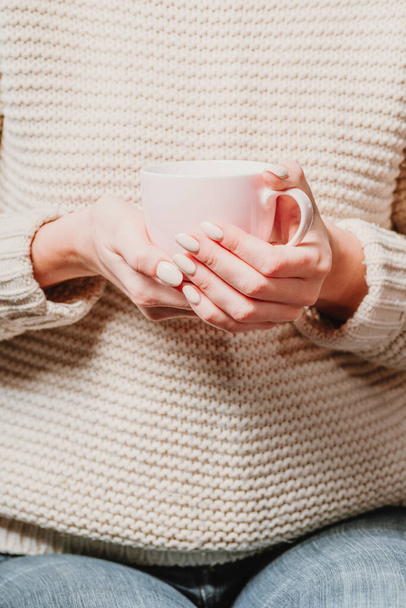 Een meisje in een beige trui houdt een kopje thee in haar handen. - Foto, afbeelding