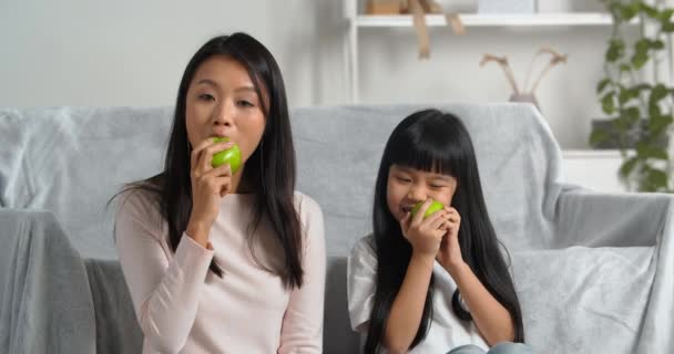 Νεαρή ασιατική οικογένεια δύο άνθρωποι μητέρα και κοριτσάκι κάθονται στο σπίτι σαλόνι τρώγοντας πράσινα ζουμερά ξινά γλυκά μήλα απολαμβάνοντας εποχιακά φρούτα τρώγοντας βιταμίνη δίαιτα σκουπίζοντας ο ένας τον άλλο μάγουλα - Πλάνα, βίντεο