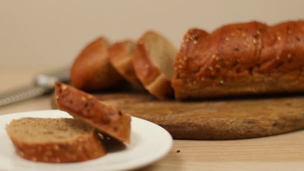 Pezzi di pane affettato di segale con semi su un piatto e una pagnotta su una tavola da cucina di legno - Filmati, video