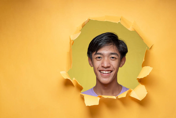 Happy Young Asian teenager man smiling poses through torn yellow paper hole, wearing purple t-shirt with a copy space or blank yellow paper. - Photo, Image