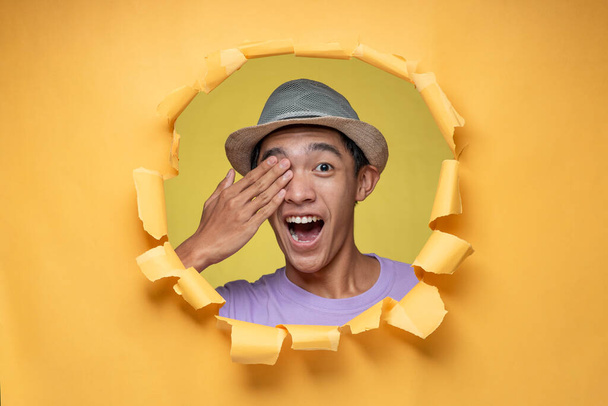 Gesto sorprendido. feliz joven asiático adolescente hombre poses a través rasgado amarillo papel agujero, cubriendo su ojo con la mano y boca abierta amplia, en amarillo aislado - Foto, Imagen