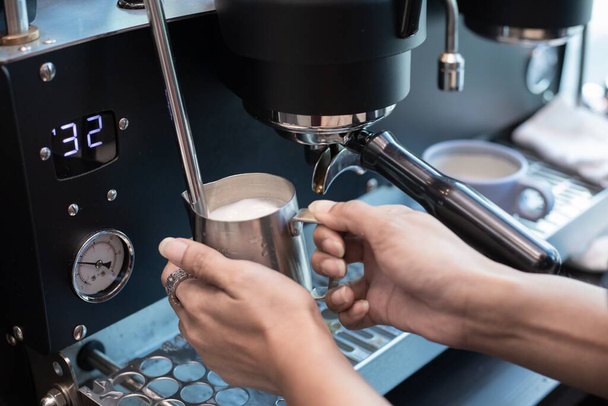 Cerrado de baristas mano sosteniendo jarra llena de leche espumosa de la máquina de café. Para hacer capuchino o latte - Foto, imagen