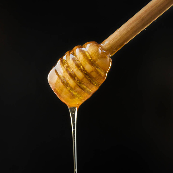 Miel que gotea de la cacerola de miel. Miel orgánica saludable que se sumerge de la cuchara de madera. Miel y cuchara de madera sobre fondo negro.   - Foto, Imagen
