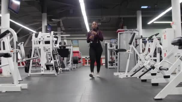 Sports training - african-american woman walks in the empty gym and drinking water from the bottle - Footage, Video