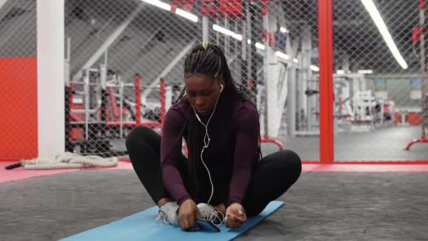Sport w siłowni - afrykańsko-amerykańska kobieta siedząca na macie do jogi w siłowni i korzystająca z telefonu - Materiał filmowy, wideo