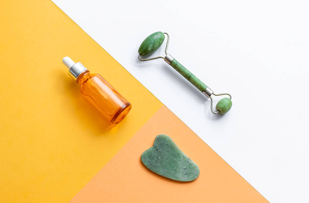 Gua sha, jade roller and face serum on geometric backgroundAnti aging skin care product for woman. Top view of vitamin c, hyaluronic acid or retinol serum, gua sha and jade roller in multi colored background - Photo, Image