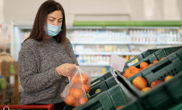 Mladá dívka v ochranné lékařské masce nakupuje v supermarketu a dává citrusové plody do sáčku v oddělení ovoce a zeleniny. Nákupní koncept v obchodě. - Fotografie, Obrázek