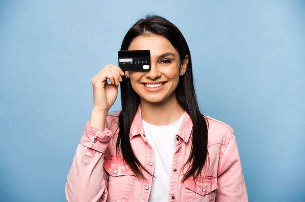 Zadowolony uśmiechnięty biały brunetka dziewczyna w stylowe ubrania trzymając kartę kredytową banku w pobliżu twarzy, stojąc na odizolowanym tle niebieski - Zdjęcie, obraz