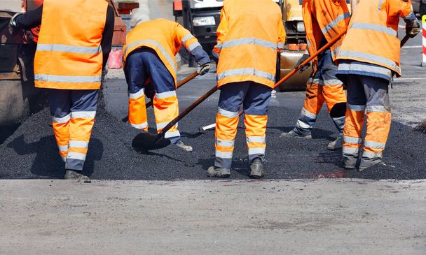 Een team van wegwerkers in oranje reflecterende uniformen repareert een stuk weg met vers asfalt. Vrije middag, kopieerruimte. - Foto, afbeelding