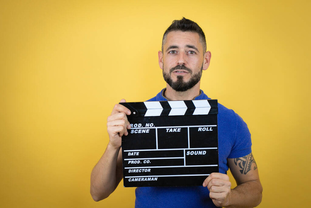Handsome man with beard wearing blue polo shirt over yellow background holding clapperboard very happy having fun - Photo, Image