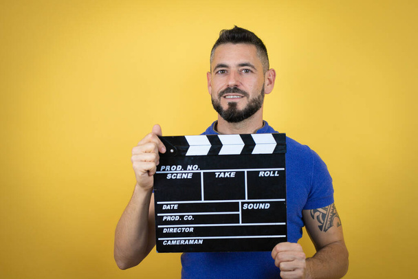 Handsome man with beard wearing blue polo shirt over yellow background holding clapperboard very happy having fun - Photo, Image