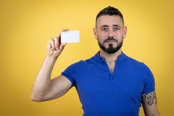 Beau homme barbu portant un polo bleu sur fond jaune souriant et tenant une carte blanche - Photo, image
