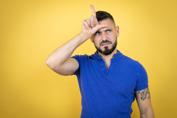 Handsome man with beard wearing blue polo shirt over yellow background making fun of people with fingers on forehead doing loser gesture mocking and insulting. - Photo, Image