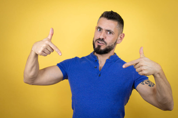 Handsome man with beard wearing blue polo shirt over yellow background looking confident with smile on face, pointing oneself with fingers proud and happy. - Photo, Image