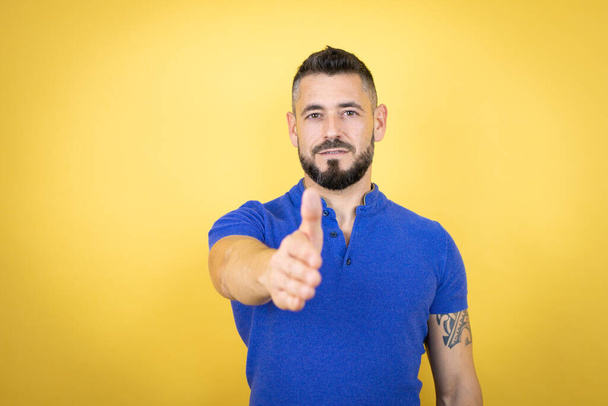 Pohledný muž s plnovousem na sobě modré polo košile přes žluté pozadí s úsměvem přátelské nabízení handshake jako pozdrav a přivítání - Fotografie, Obrázek