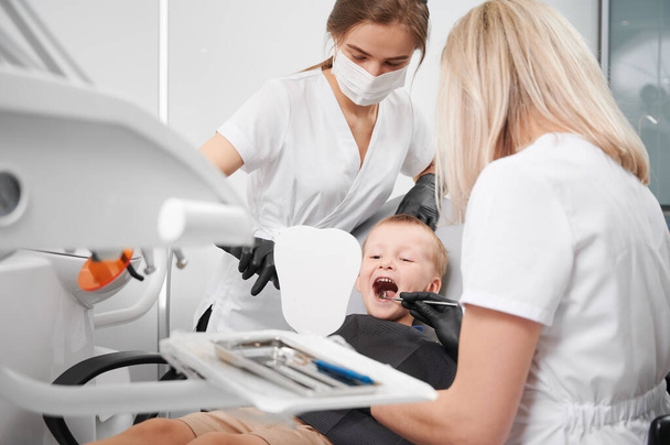 Το αγόρι κάθεται στην οδοντιατρική καρέκλα ενώ δύο γυναίκες οδοντίατροι ελέγχουν παιδικά δόντια. Γυναίκα στοματολόγος εξέταση μικρά δόντια αγόρι με οδοντιατρικό όργανο, ενώ βοηθός στην ιατρική μάσκα που δείχνει στον καθρέφτη. - Φωτογραφία, εικόνα