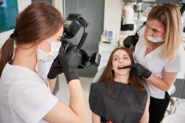 Dentista examinando dentes de mulher enquanto assistente tirando foto com câmera profissional. Médica mulher fotografando paciente e dentista em consultório odontológico. Conceito de odontologia, cuidados dentários, fotografia. - Foto, Imagem
